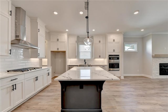 kitchen with wall chimney exhaust hood, decorative light fixtures, light stone countertops, appliances with stainless steel finishes, and a center island