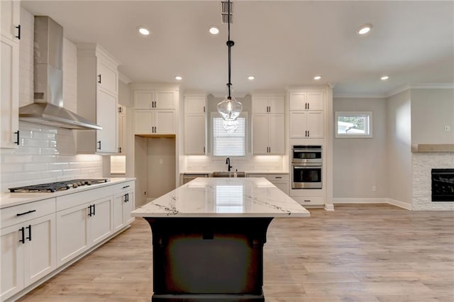 kitchen featuring an island with sink, wall chimney exhaust hood, crown molding, appliances with stainless steel finishes, and decorative light fixtures