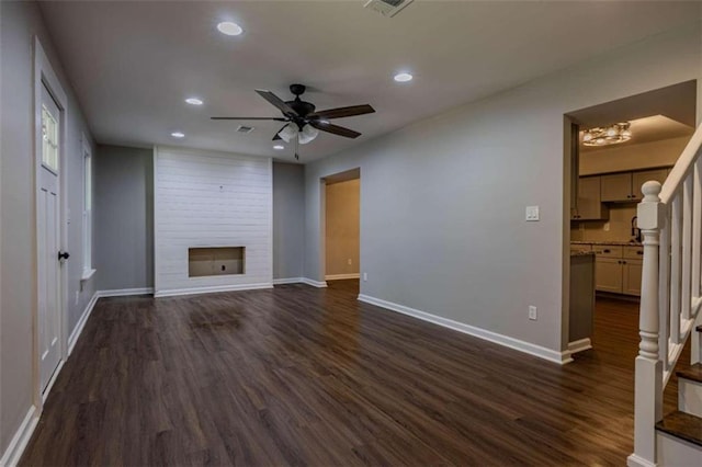 unfurnished living room featuring ceiling fan with notable chandelier, a large fireplace, and dark wood-type flooring