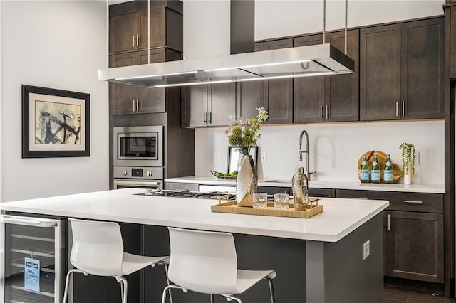 kitchen featuring a center island with sink, wall chimney exhaust hood, appliances with stainless steel finishes, dark brown cabinets, and beverage cooler