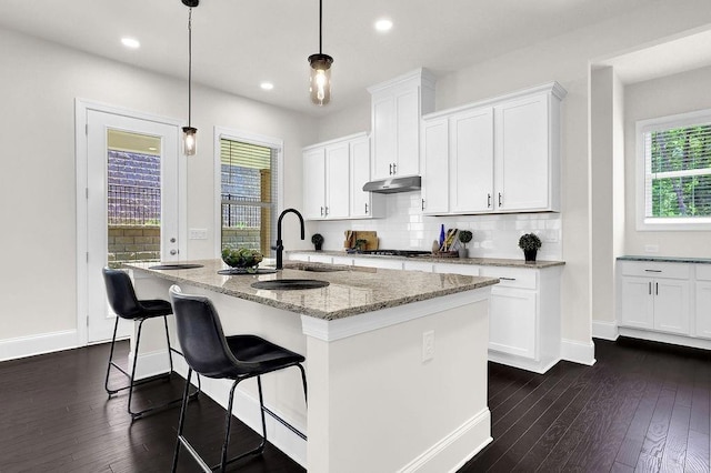 kitchen with light stone countertops, hanging light fixtures, decorative backsplash, white cabinets, and a center island with sink