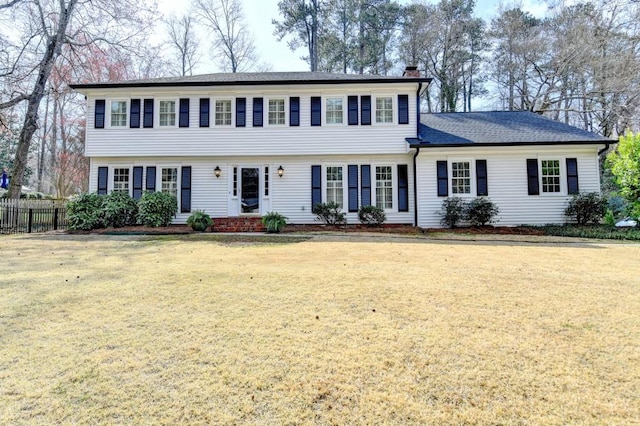 colonial house featuring a front yard, fence, roof with shingles, and a chimney