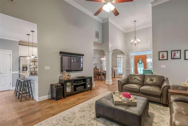 living room with a towering ceiling, crown molding, decorative columns, ceiling fan with notable chandelier, and light wood-type flooring