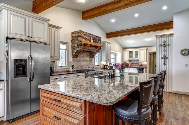 kitchen with vaulted ceiling with beams, appliances with stainless steel finishes, an island with sink, light wood-type flooring, and tasteful backsplash