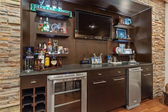 bar with dark brown cabinets, stainless steel counters, light wood-type flooring, and beverage cooler