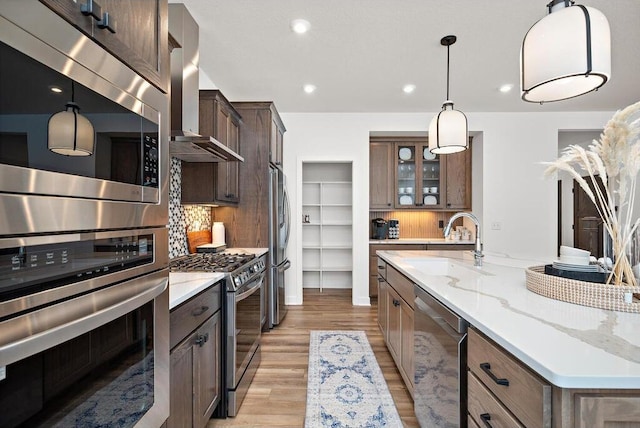 kitchen with backsplash, appliances with stainless steel finishes, an island with sink, light wood-type flooring, and wall chimney range hood