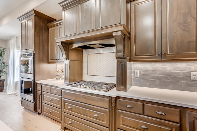 kitchen with exhaust hood, backsplash, light hardwood / wood-style floors, and appliances with stainless steel finishes