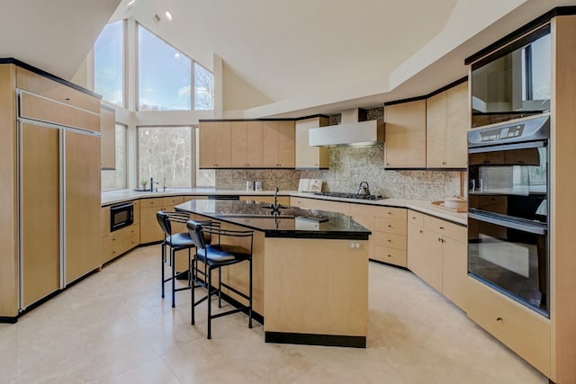 kitchen featuring light brown cabinets, high vaulted ceiling, black appliances, a center island with sink, and wall chimney range hood