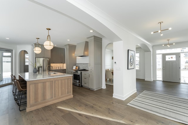 kitchen featuring stainless steel appliances, arched walkways, a kitchen island with sink, and wall chimney range hood