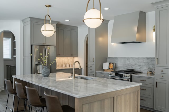 kitchen with arched walkways, a kitchen island with sink, ventilation hood, appliances with stainless steel finishes, and light stone countertops