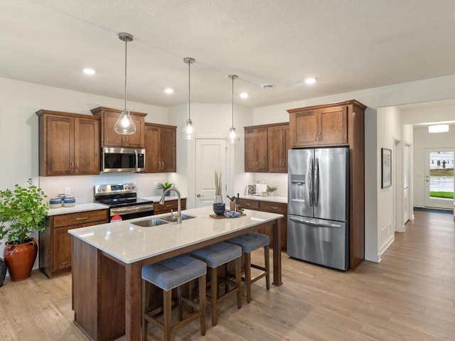 kitchen with an island with sink, sink, decorative light fixtures, light wood-type flooring, and appliances with stainless steel finishes