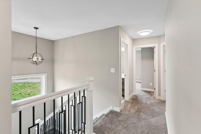 hallway featuring baseboards, dark colored carpet, an upstairs landing, and a notable chandelier