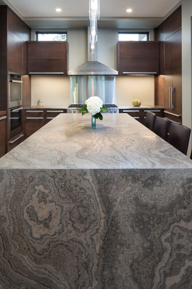 kitchen with double oven, wall chimney range hood, a kitchen island, and a kitchen bar