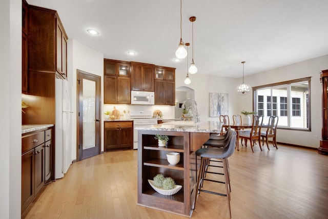 kitchen featuring light stone countertops, hanging light fixtures, light hardwood / wood-style flooring, an island with sink, and white appliances