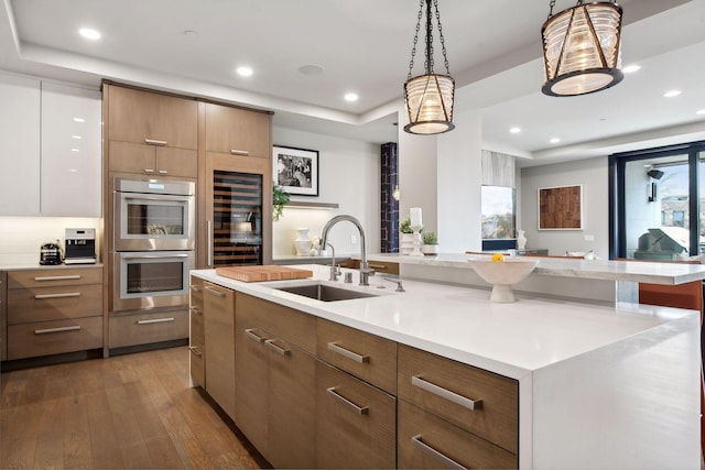 kitchen featuring an island with sink, dark hardwood / wood-style floors, double oven, sink, and hanging light fixtures