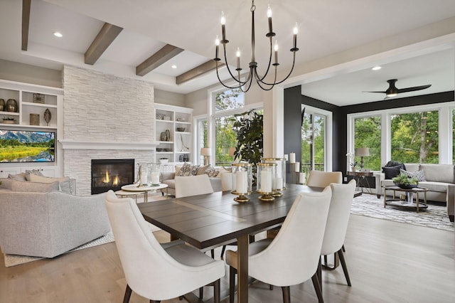 dining area featuring beam ceiling, ceiling fan with notable chandelier, a stone fireplace, built in shelves, and light wood-type flooring