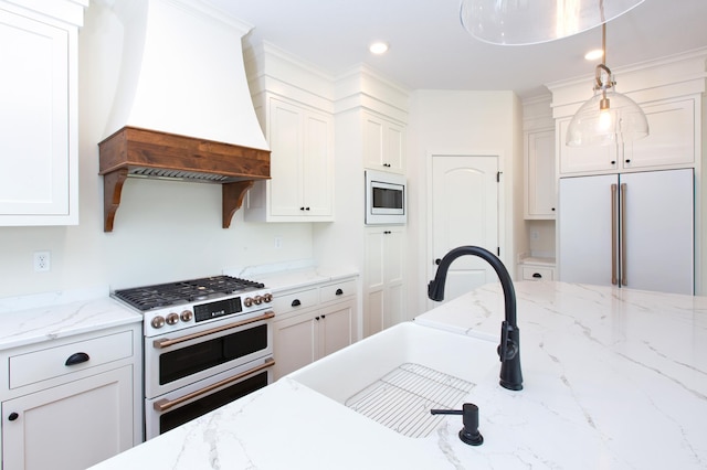 kitchen with custom range hood, light stone counters, decorative light fixtures, built in appliances, and white cabinetry