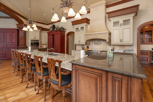 kitchen featuring premium range hood, an inviting chandelier, backsplash, a kitchen island with sink, and sink