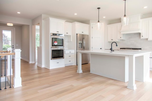 kitchen featuring a kitchen island with sink, light wood-type flooring, decorative light fixtures, stainless steel appliances, and white cabinetry