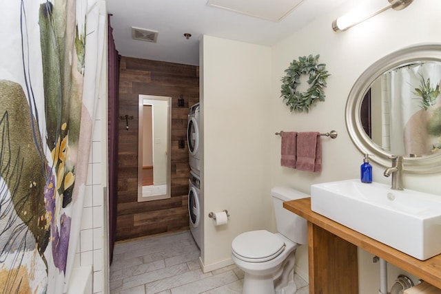 bathroom with toilet, wooden walls, stacked washer / drying machine, tile flooring, and vanity