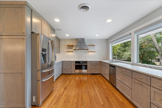 kitchen featuring appliances with stainless steel finishes, tasteful backsplash, sink, wall chimney range hood, and light hardwood / wood-style flooring