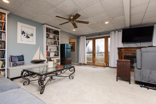 living room featuring light carpet, a drop ceiling, built in shelves, ceiling fan, and a fireplace