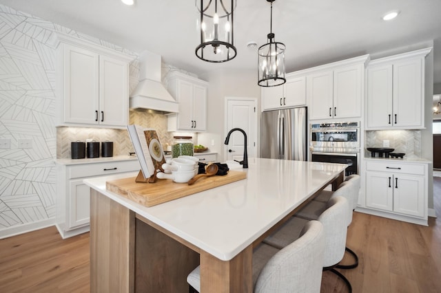 kitchen featuring appliances with stainless steel finishes, tasteful backsplash, custom exhaust hood, an island with sink, and light wood-type flooring