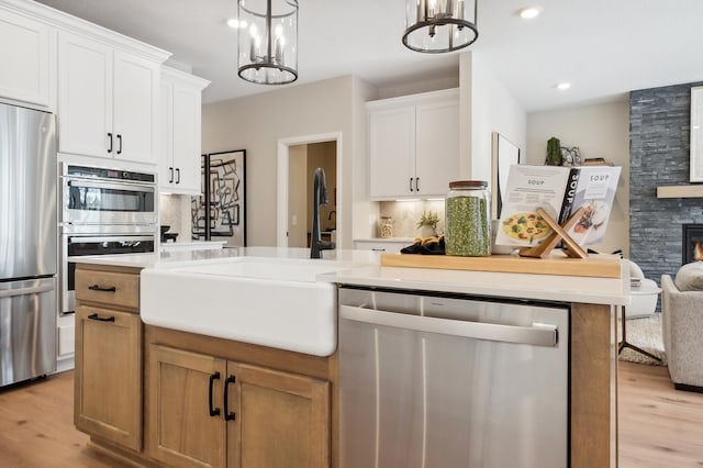 kitchen featuring appliances with stainless steel finishes, a large fireplace, white cabinetry, a notable chandelier, and light wood-type flooring