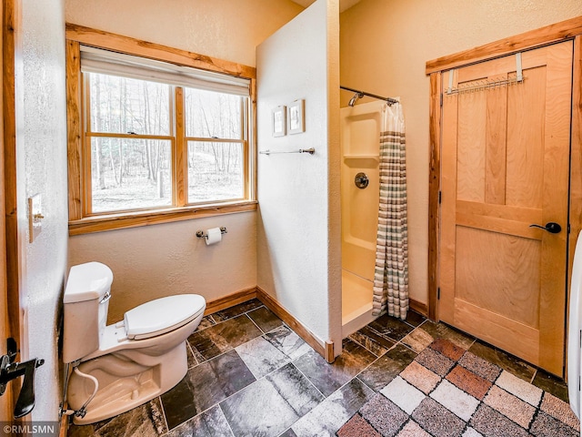 bathroom with a shower with curtain, stone tile flooring, baseboards, toilet, and a textured wall