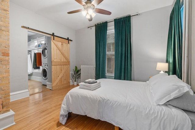 bedroom featuring ceiling fan, a barn door, wood-type flooring, radiator, and stacked washer and dryer