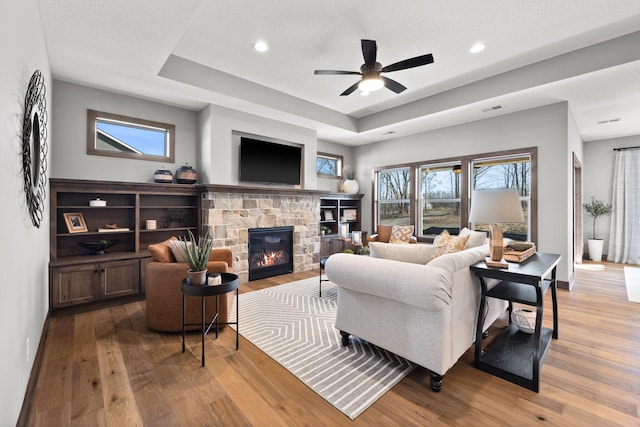 living room with ceiling fan, a stone fireplace, a textured ceiling, and light hardwood / wood-style flooring