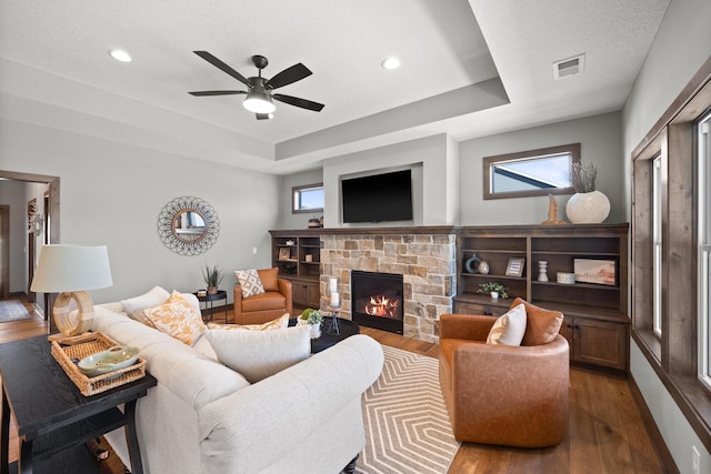 living room featuring ceiling fan, a stone fireplace, a raised ceiling, and hardwood / wood-style floors