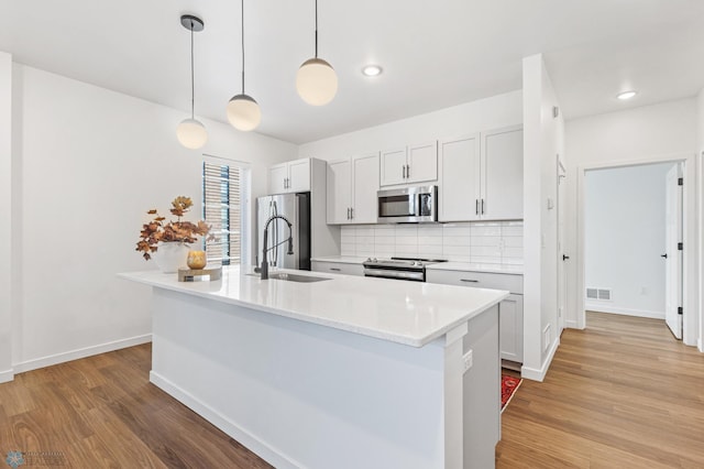 kitchen with stainless steel appliances, a center island with sink, hanging light fixtures, and light wood-type flooring