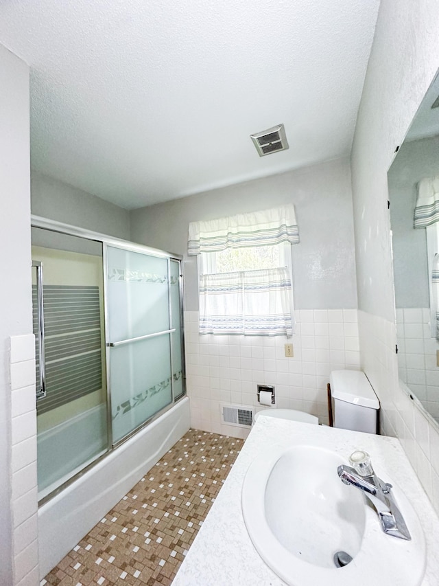 full bathroom with a textured ceiling, tile walls, shower / bath combination with glass door, toilet, and sink