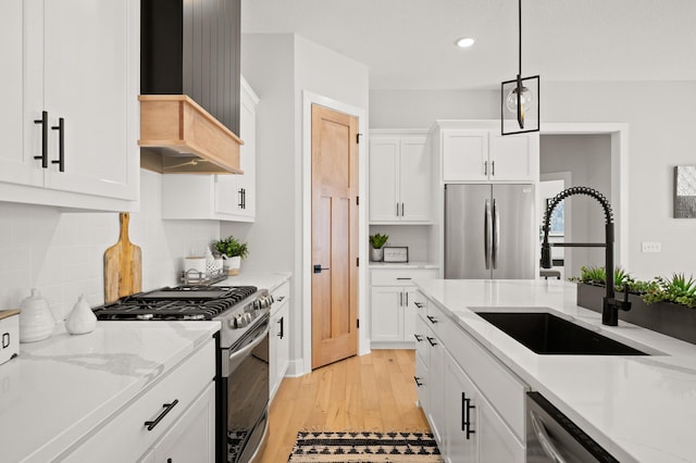 kitchen with appliances with stainless steel finishes, hanging light fixtures, wall chimney exhaust hood, sink, and light wood-type flooring