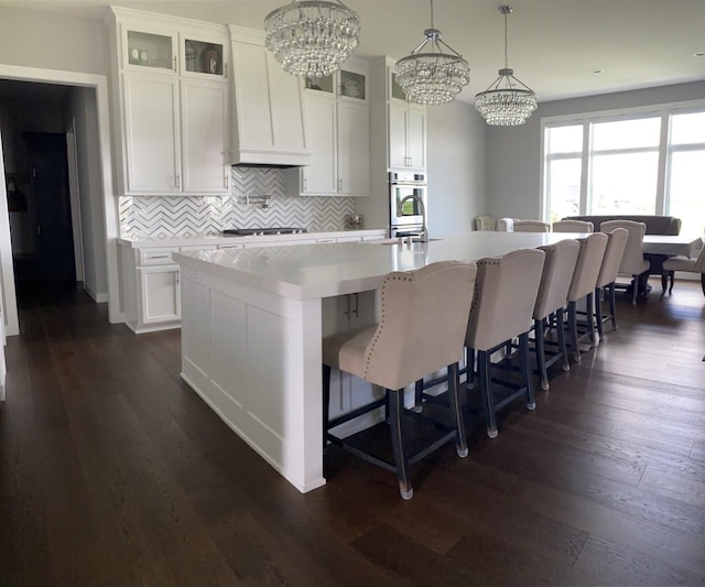 kitchen featuring decorative backsplash, dark wood-type flooring, decorative light fixtures, a center island with sink, and white cabinets