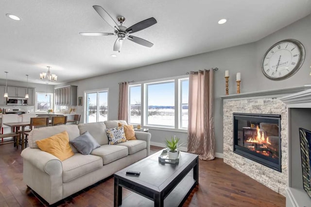 living room with ceiling fan with notable chandelier, a stone fireplace, and dark wood-type flooring