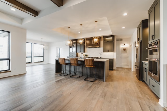 kitchen featuring custom range hood, pendant lighting, beamed ceiling, light hardwood / wood-style floors, and an island with sink