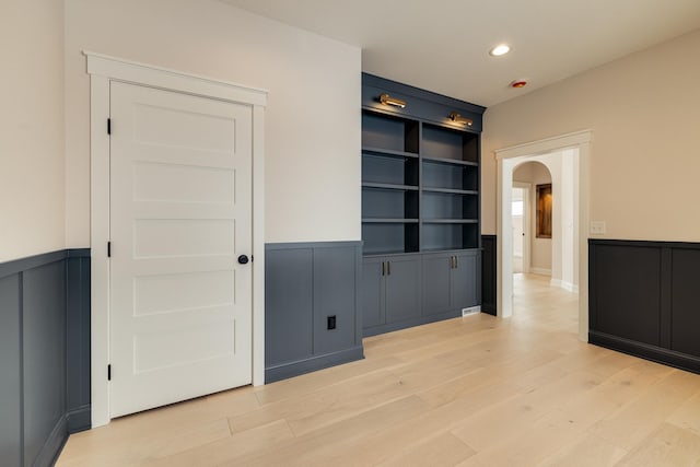 empty room featuring arched walkways, built in shelves, light wood-style flooring, recessed lighting, and wainscoting