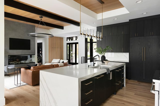 kitchen with dark cabinets, a sink, light wood-type flooring, light countertops, and beamed ceiling