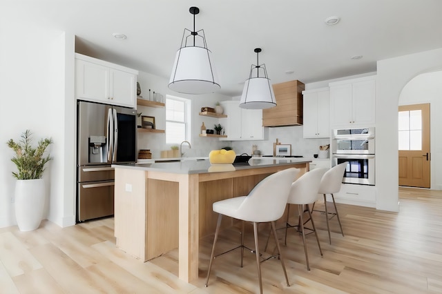 kitchen with open shelves, custom exhaust hood, an island with sink, arched walkways, and stainless steel appliances