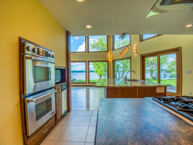 kitchen featuring stainless steel double oven, a textured ceiling, an island with sink, sink, and light tile patterned flooring