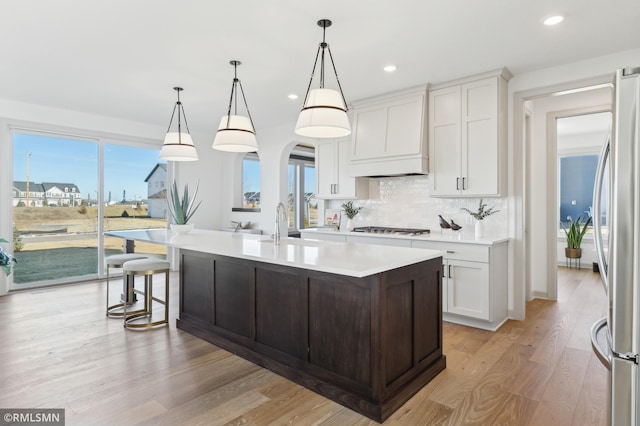 kitchen featuring decorative light fixtures, a kitchen island with sink, appliances with stainless steel finishes, custom range hood, and white cabinets