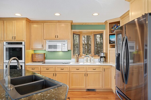 kitchen with stainless steel fridge, white microwave, multiple ovens, black electric cooktop, and light brown cabinets