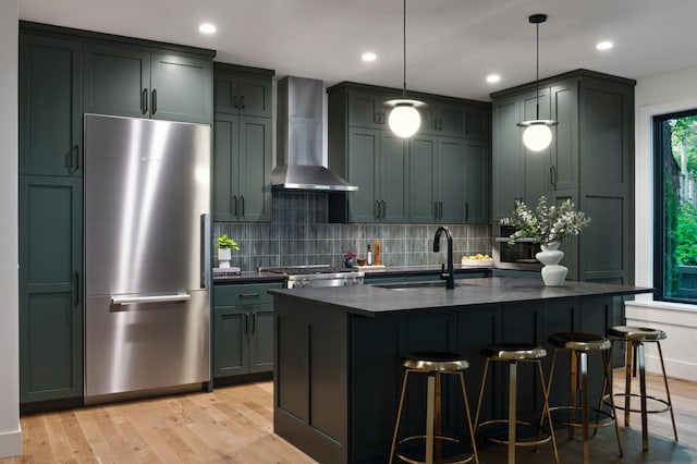 kitchen with pendant lighting, stainless steel refrigerator, an island with sink, sink, and wall chimney exhaust hood