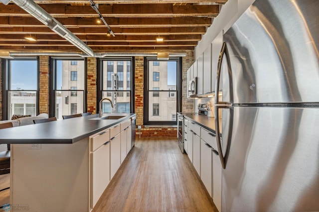 kitchen with sink, an island with sink, appliances with stainless steel finishes, white cabinetry, and brick wall