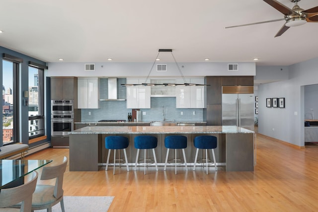 kitchen with stainless steel appliances, a sink, wall chimney range hood, ceiling fan, and backsplash