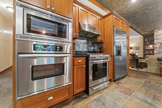kitchen with under cabinet range hood, brown cabinets, appliances with stainless steel finishes, and stone finish flooring