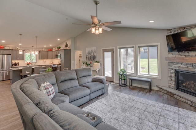 living room featuring high vaulted ceiling, light hardwood / wood-style flooring, ceiling fan, and a stone fireplace