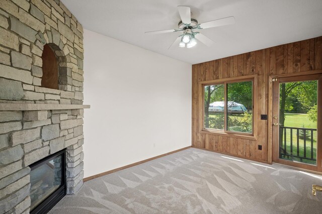 unfurnished living room with wood walls, a stone fireplace, ceiling fan, and carpet floors